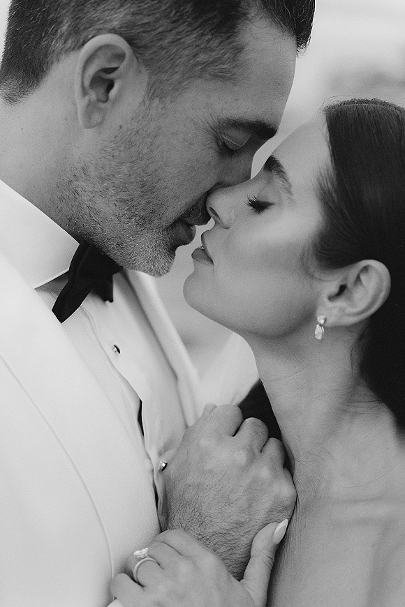 Wedding kiss portrait in black and white, bride and groom nose-to-nose with her ringed hand on his white tuxedo lapel, blurred backdrop