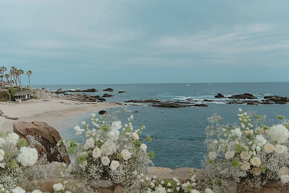 Ocean view ceremony with white rose and baby's breath floral arrangements and greenery on a stone ledge above a rocky coastline and beach