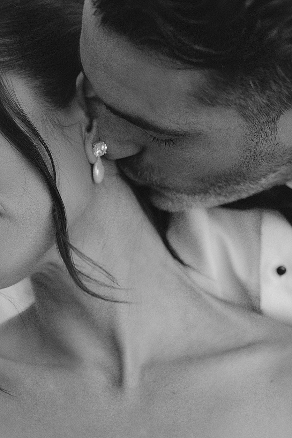 Wedding kiss portrait of groom kissing bride’s cheek in close up, her pearl drop earrings visible beside his bow tie against a neutral backdrop