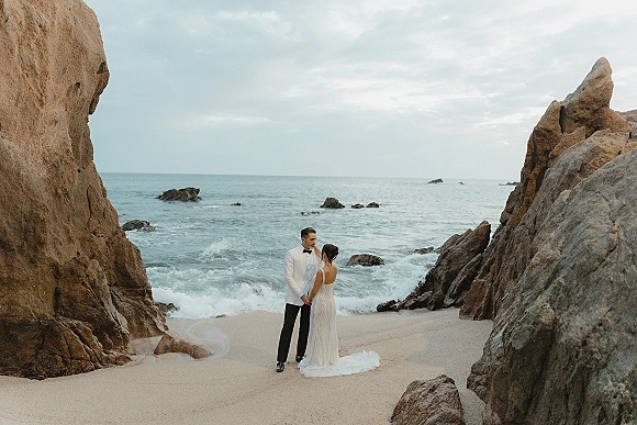 Couple portrait of newlyweds holding hands on a rocky beach, bride’s veil blowing and dress train on sand with waves and cliffs behind