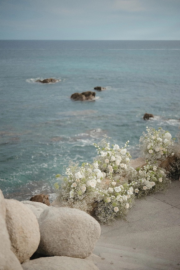 Ceremony aisle florals with wedding aisle flowers in white roses, baby's breath, and greenery lining stone steps beside an ocean shoreline