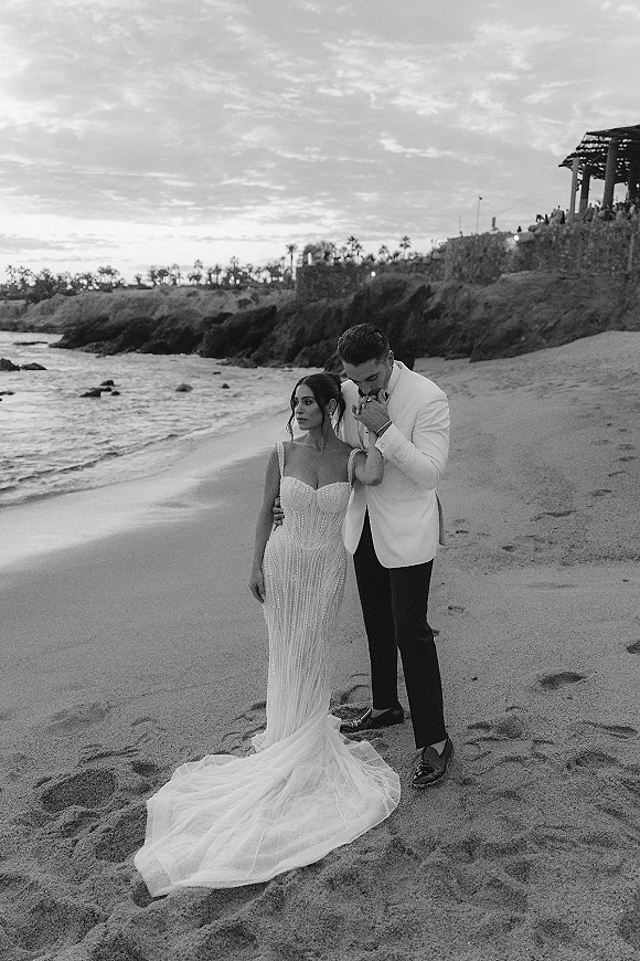 Couple portrait on a beach as the groom kisses the bride’s hand beside ocean waves, her beaded gown and long train flowing in the wind