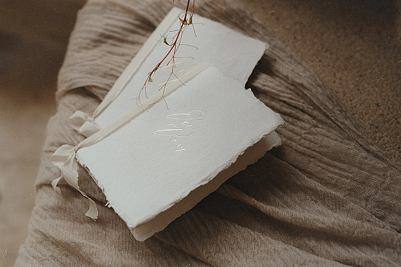 Vow books with handmade deckled edges and silk ribbon ties, calligraphy lettering and a dried floral stem on linen fabric over stone surface