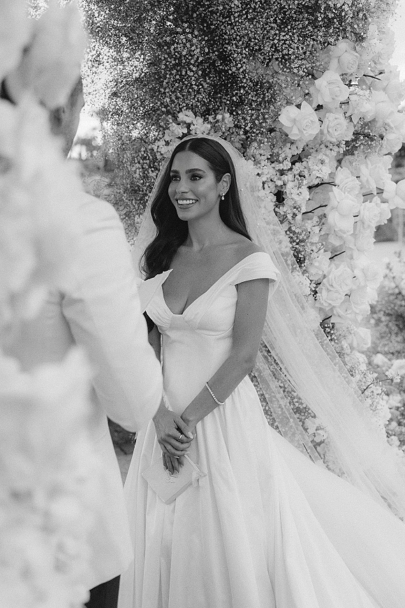 Ceremony moment as the bride at the altar smiles at the groom facing bride, holding a vow book beneath a rose arch in a garden