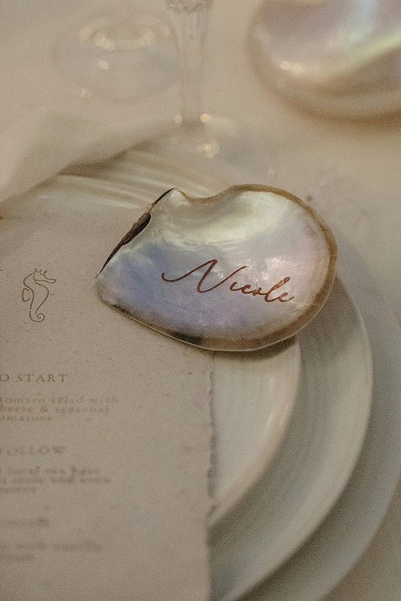 Place card on a gold-edged oyster shell with calligraphy beside a dinner plate, wine glass, and menu on a white tablecloth