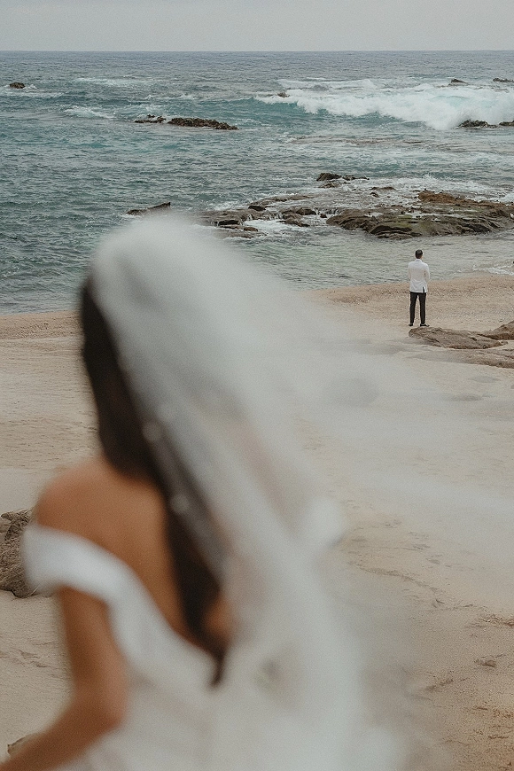 Wedding first look on a beach as the bride approaches the groom, her veil blowing in the wind near ocean waves and rocky shoreline