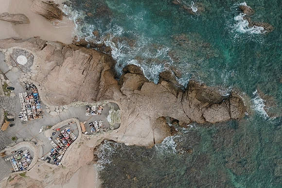 Cliffside wedding ceremony with oceanfront ceremony setup, white chair rows and floral aisle on a stone patio above rocky shoreline waves
