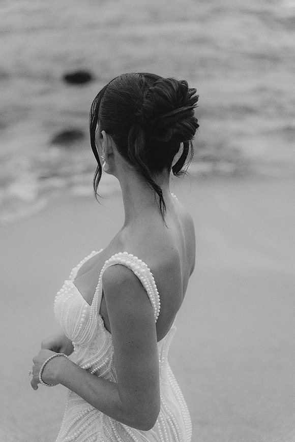 Bridal portrait in black and white of a bride looking away, showing a strapless pearl-beaded gown and updo with ocean water behind her