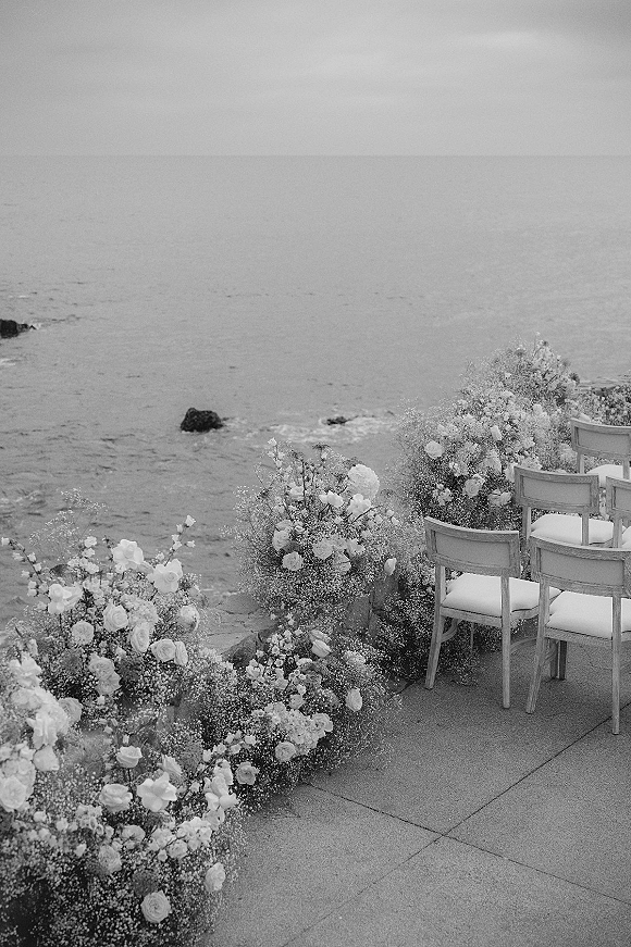 Ceremony aisle decor with white rose and baby's breath floral clusters lining chair rows on a stone patio overlooking the ocean horizon