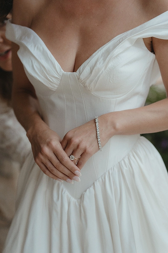 Wedding dress detail of a bride adjusting an off the shoulder wedding dress corset bodice, showing engagement ring, tennis bracelet, manicured nails against blurred greenery
