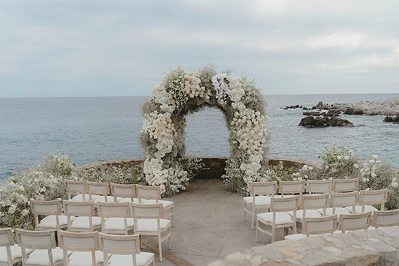 Ceremony setup for an oceanfront wedding ceremony with a floral arch of white flowers and baby's breath on a stone terrace by rocky coastline