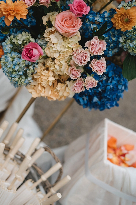 Wedding bouquet with blue hydrangea bouquet tones, pink roses and greenery beside a wicker basket and wooden fans on stone ground