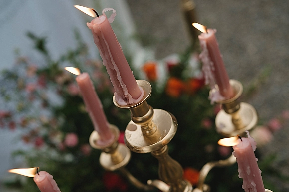 Wedding candles in a brass candelabra centerpiece with pink taper flames, wax drips, and greenery beside a blurred outdoor floral arrangement