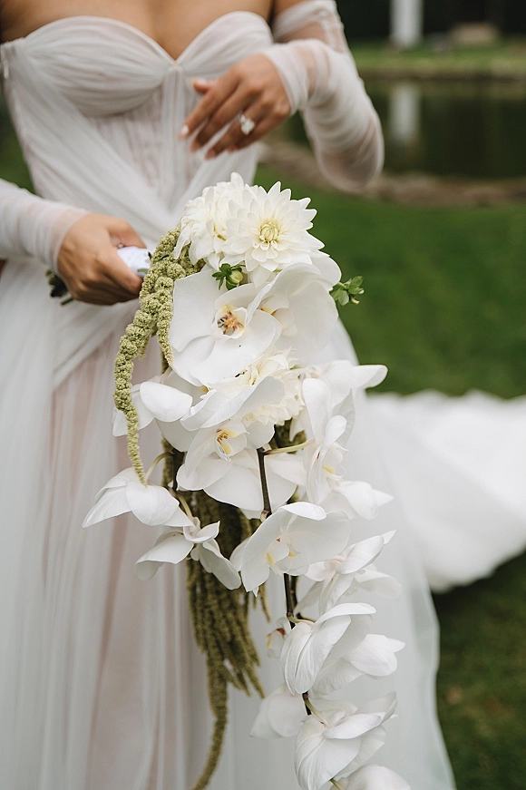 Bridal bouquet of cascading orchid blooms with white dahlia and green hanging amaranthus held against an off-shoulder gown on a lawn