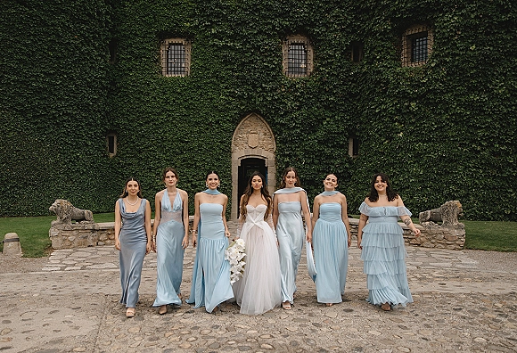Bride with bridesmaids in blue dresses walking beside an ivy-covered stone building, holding a white orchid bouquet and wearing pearls