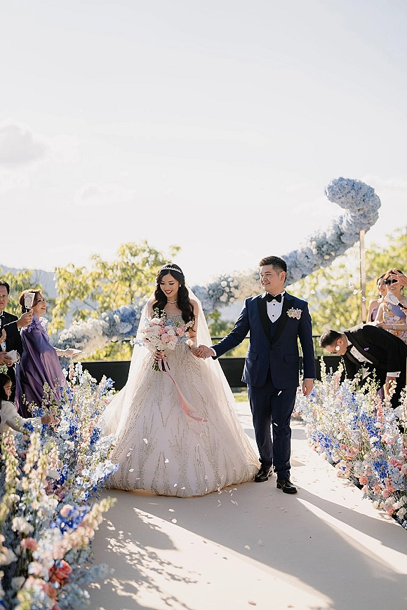Wedding recessional as bride and groom walk the aisle holding hands, bride with bouquet and tiara veil, petals tossed in garden setting