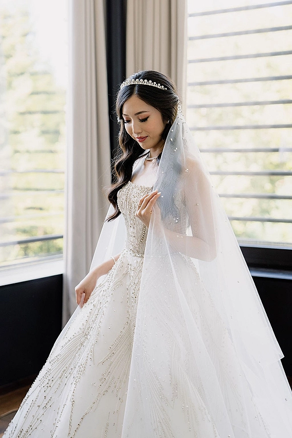 Bridal portrait of a tiara bride look in a beaded wedding dress, holding her cathedral veil by a large window with blinds in natural light
