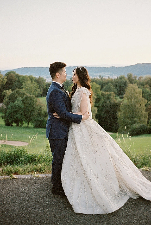 Couple portrait of bride and groom embrace, her beaded gown and veil flowing in wind on a hilltop path with mountains behind