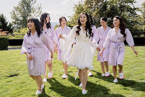 Bridesmaids getting ready in bridal party robes, walking together in lavender and white robes on a garden lawn with trees and railing
