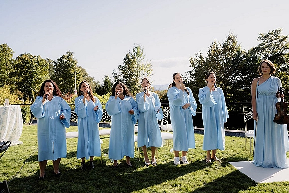Wedding ceremony musicians in blue choir robes sing into microphones beside a violinist on an outdoor lawn with trees and sky