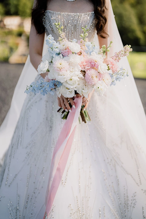 Bridal bouquet of soft pastel wedding bouquet blooms tied with a pink ribbon, held against a strapless beaded gown and veil on a garden path