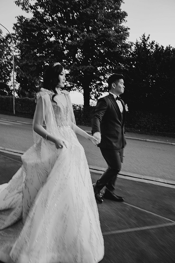 Couple portrait of bride and groom walking hand in hand, her long veil trailing as they stroll along a tree-lined street sidewalk