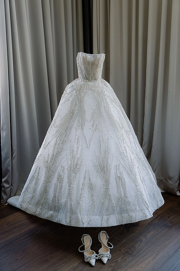 Wedding dress in a strapless ball gown silhouette with beaded embellishments, hanging by curtains above bridal heels on a wood floor