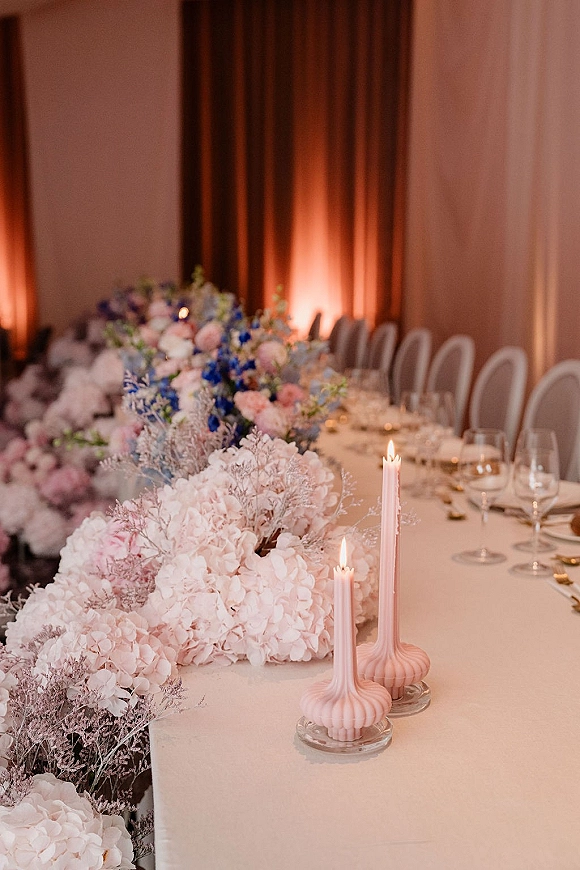 Reception tablescape with wedding head table decor, hydrangea floral runner, pink taper candles in glass holders, and place settings in a draped, uplit banquet room