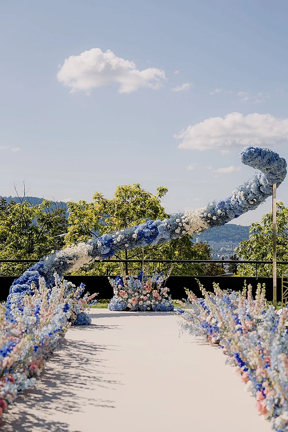Ceremony aisle decor with a wedding floral arch and pastel blue, white, and pink aisle flowers lining a runner on an outdoor terrace with hills