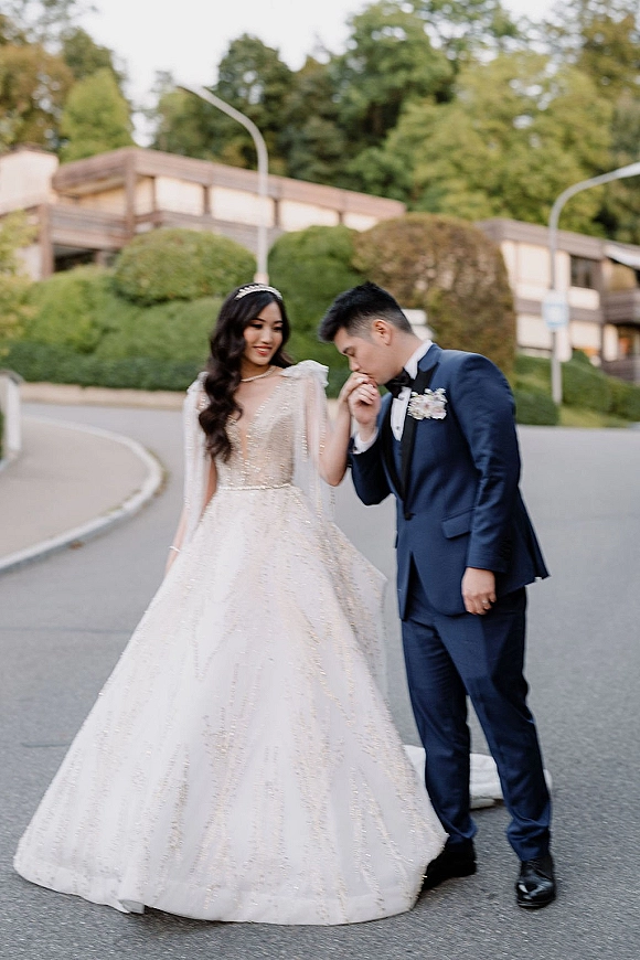 Couple portrait of bride and groom walking as he kisses her hand, her veil and tiara flowing, on a tree-lined road with streetlights