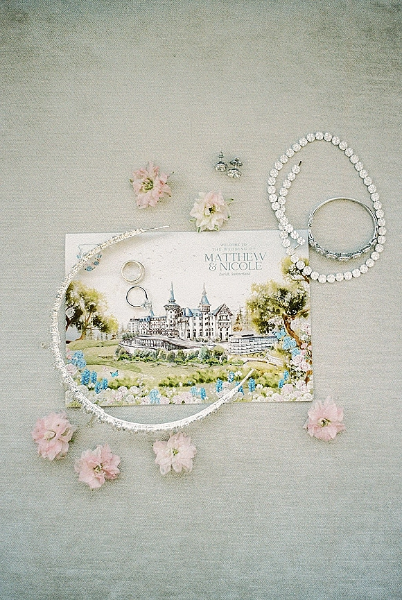 Invitation suite wedding invitation flat lay with engagement ring, band and crystal jewelry beside pink flowers on a gray fabric backdrop