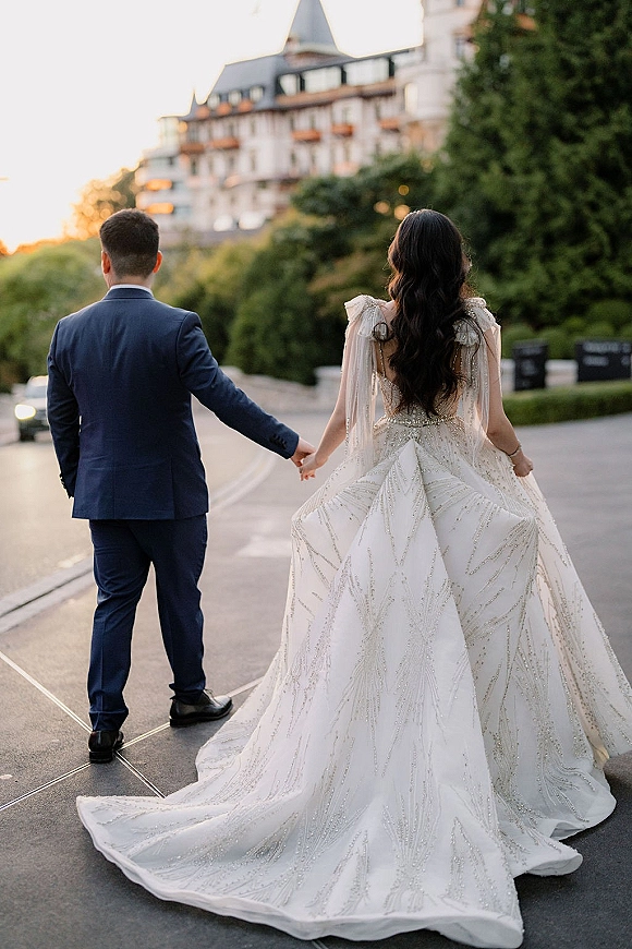 Couple portrait of bride and groom holding hands, walking away on a road at sunset, her beaded train flowing behind near streetlights