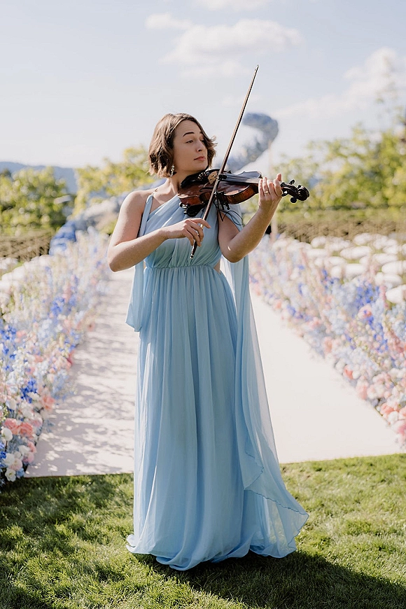 Wedding violinist in a light blue bridesmaid dress playing with bow and violin along a flower-lined aisle runner on a mountain lawn