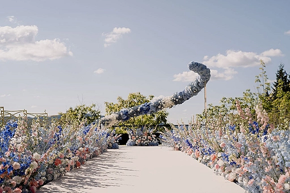 Ceremony aisle decor with an outdoor ceremony aisle of blue, white, and blush ground florals, scattered petals, and a floral arch on a terrace under clouds
