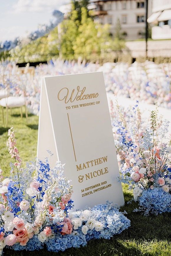 Wedding welcome sign on clear acrylic with gold lettering, framed by hydrangea and rose florals on a lawn with mountain view behind