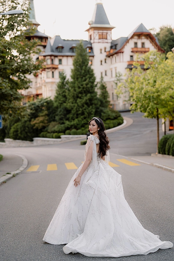 Bridal portrait of a bride walking away, glancing over her shoulder in a beaded ball gown with long train and tiara outside a castle-like hotel