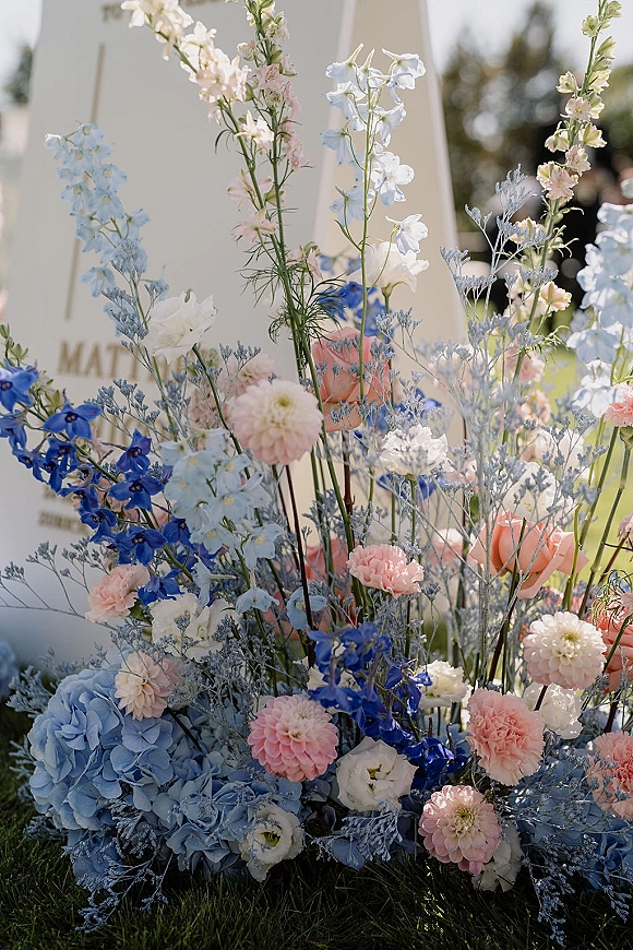 Wedding welcome sign with acrylic welcome sign styling on an easel, framed by blue and blush florals on a sunlit lawn with trees behind