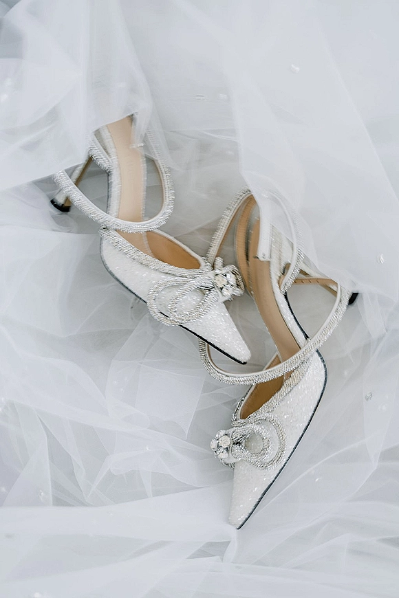 Bridal shoes with sparkly wedding heels featuring pointed toes, ankle straps, rhinestone trim, and a crystal bow on white fabric