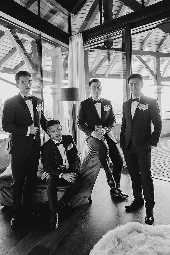 Groomsmen portrait of men in black tuxedos with bow ties, holding drink glasses and a camera by glass wall windows and beams