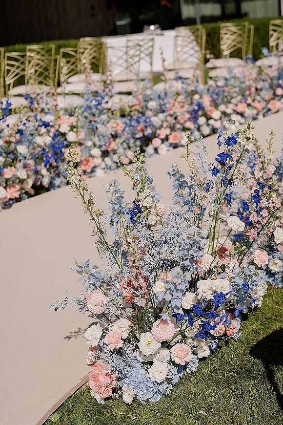 Ceremony aisle flowers and wedding aisle florals arranged in pastel blue and pink clusters along a grass lawn beside gold Chiavari chairs