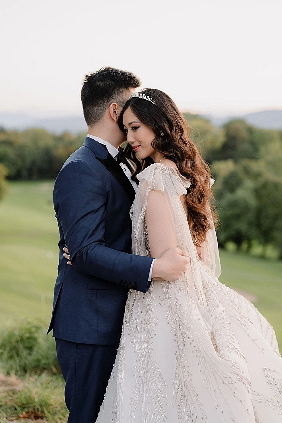 Couple portrait of bride and groom embrace as he whispers to her, bride in beaded gown and tiara in a green field with rolling hills
