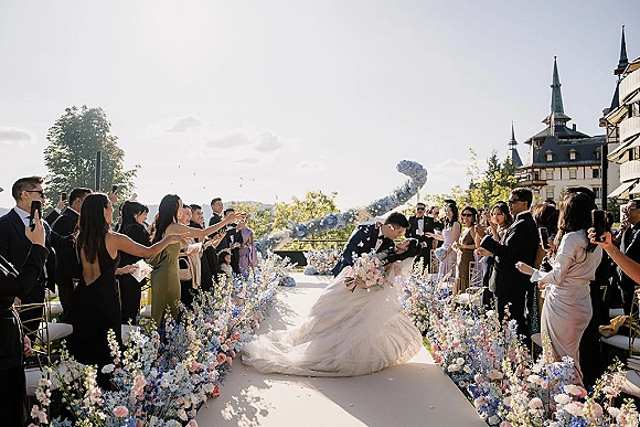 Ceremony kiss moment as groom dips bride in a strapless ball gown, guests tossing flower petals on a floral-lined castle terrace aisle