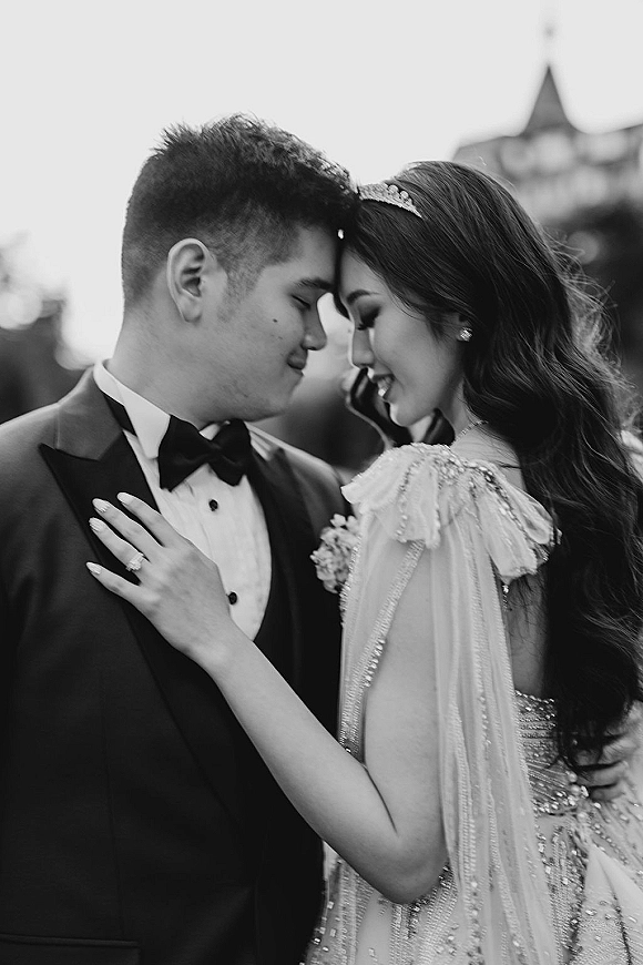 Couple portrait in a black and white wedding portrait, bride and groom forehead touch in embrace, her tiara and ring visible outdoors near a blurred building