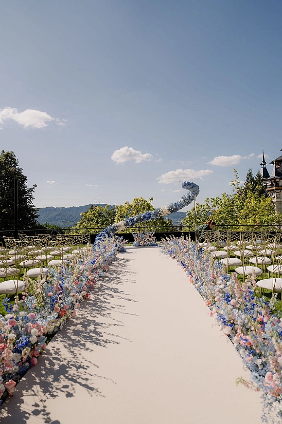 Ceremony aisle decor with a white runner and pastel blue and pink florals leading to a floral arch on an outdoor terrace with mountain views