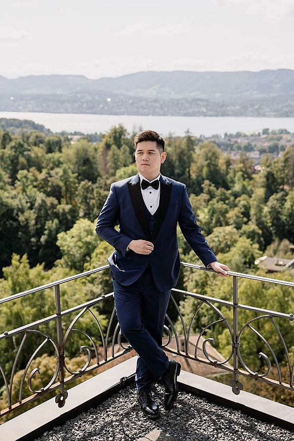 Groom portrait in a navy tuxedo, leaning on a balcony railing with mountain and lake view behind, forest trees and sky beyond