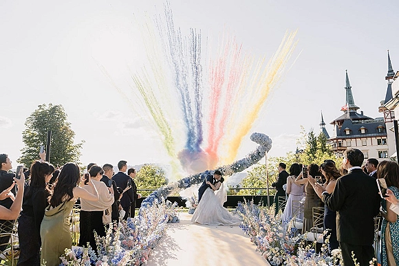 Wedding kiss moment as the bride and groom kiss under a floral arch on an outdoor castle terrace, rainbow smoke bombs rising behind them
