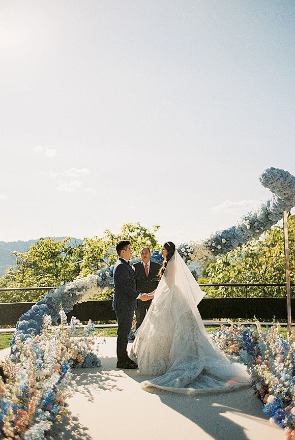 Wedding vows as bride and groom hold hands under a blue floral arch on an outdoor terrace, with aisle flowers and hills beyond