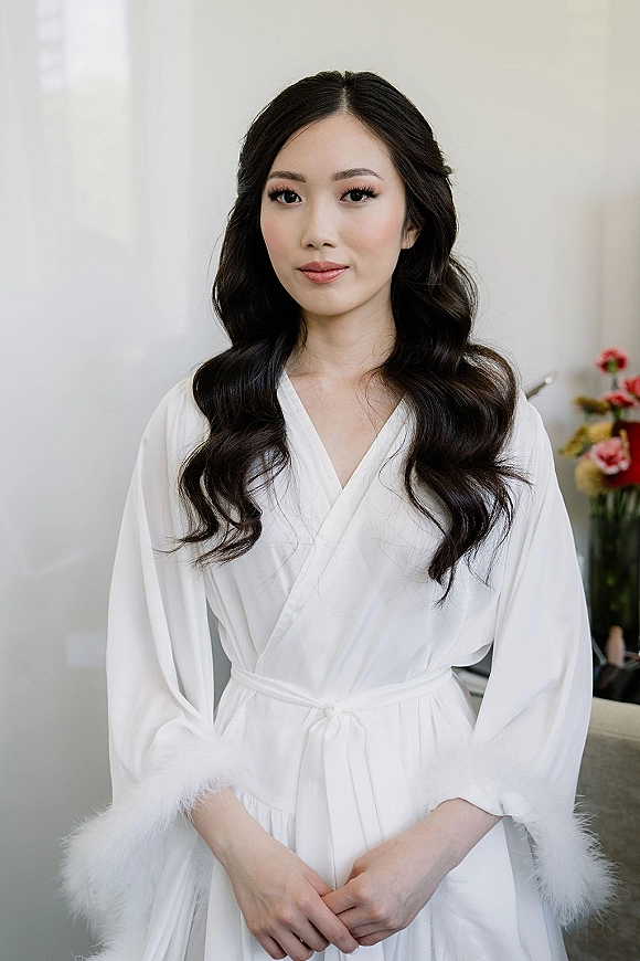 Bridal portrait of a bride getting ready in a white satin robe with feather cuffs, standing by a window in an indoor room with florals
