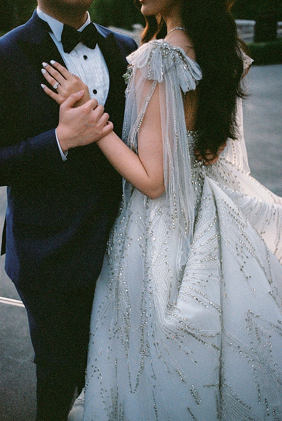 Couple portrait of bride holding groom’s arm, showing her engagement ring and beaded off-shoulder gown on a leafy walkway