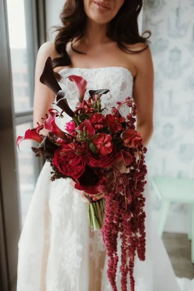 Bridal bouquet of deep red roses and calla lilies in a cascading shape, held against a strapless lace dress in window light